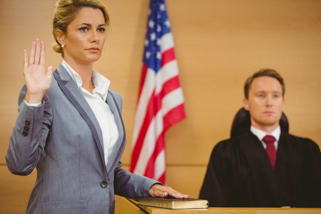 A blonde woman standing in court with her hand on the bible swearing to tell the truth, there is a judge in the background. Representing how one can benefit from calling a Portland criminal defense attorney.