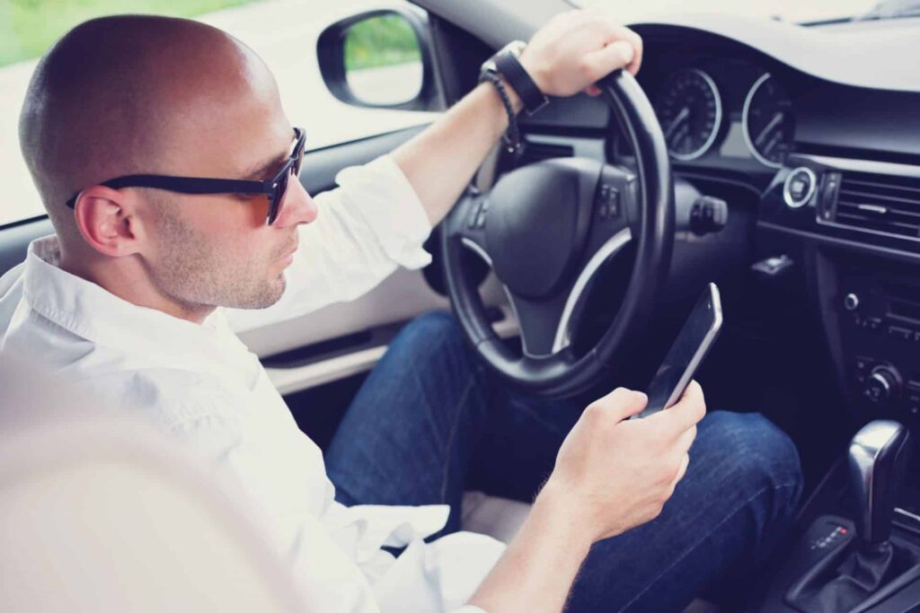 What is driving to endanger Offenses in Maine portrayed by a man inside a car, sitting on the driver's seat holding the wheel with one hand and his phone with the other, representing how one can benefit from calling a Portland criminal defense lawyer.